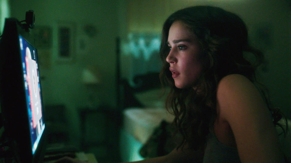 A young woman intently looks at a computer screen while sitting in a dimly lit room