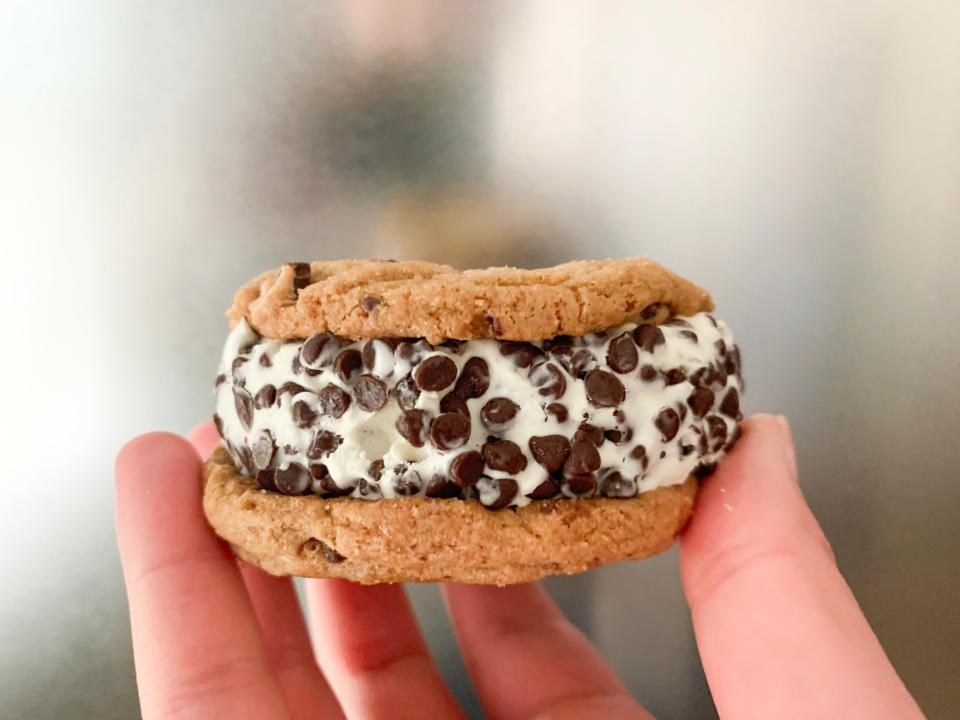 A hand holds an ice-cream sandwich so the side of it is visible. The ice-cream sandwich is made with chocolate-chip cookies and has a vanilla filled with chocolate chips on the edges