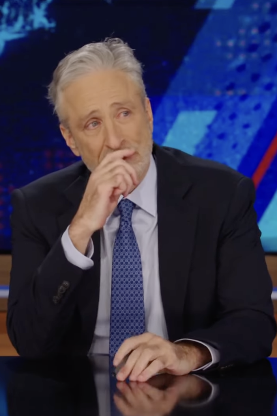 Jon Stewart seated at The Daily Show desk, looking pensive with his hand on his chin