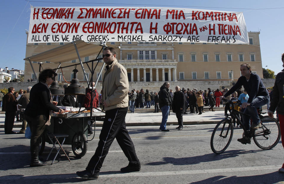 A cyclist and a street vendor are seen under a banner which reads ''National consensus is a trick, poverty and hunger have no nationality'' during a peaceful protest outside the Greek Parliament in Athens, Sunday, Feb. 19, 2012. Tensions between Athens and other European capitals hit new highs this week as eurozone ministers delayed to next Monday a decision on a bailout agreement and demanded more commitments from Greece. (AP Photo/Thanassis Stavrakis)