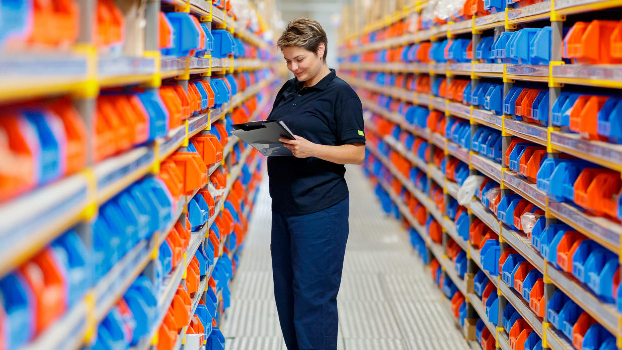 Woman with checklist examining red and blue trays on shelves in the aisle of warehouse.