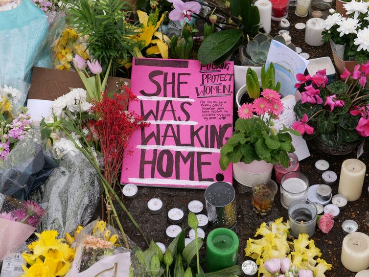 A pink sign with the handwritten message 'She was walking home', on the ground surrounded by flowers and candles