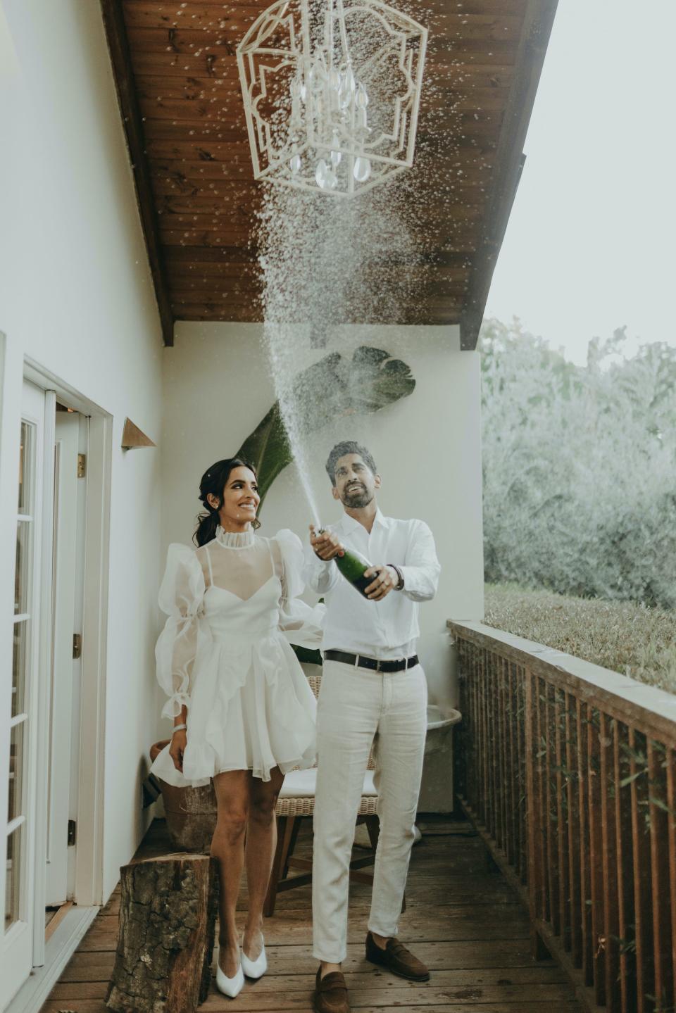 A bride and groom pop champagne on a porch on their wedding day.