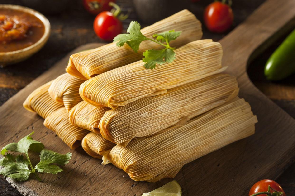 Tamales on a wooden cutting board, with cilantro, cut limes and cherry tomatoes