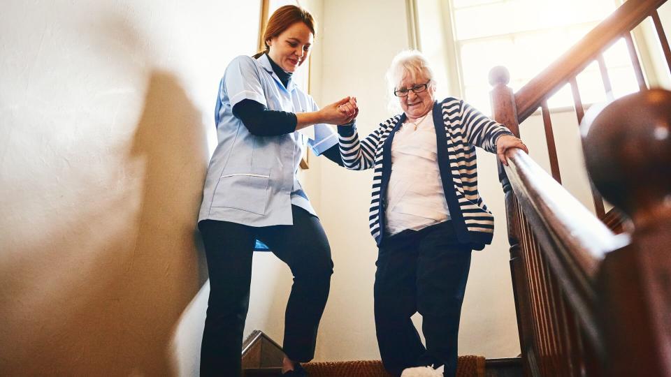 Young female caregiver helping senior woman walking down stairs at home.