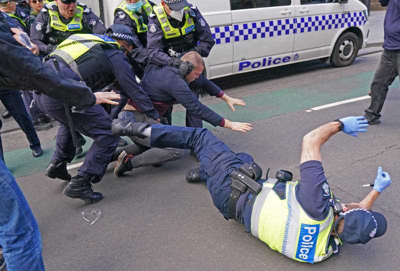 A police officer falls to the ground as officers try to detain a man while protesters gather outside Parliament House in Melbourne.