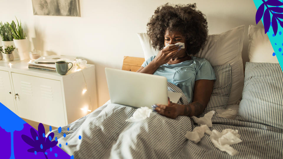 A woman sits in bed propped up against the pillows with a pained expression and dozens of discarded tissues around her, as she looks at her laptop. 