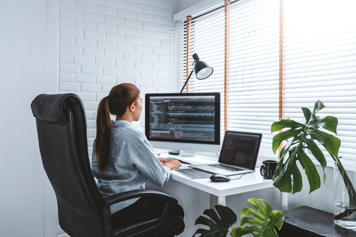 Female information security analyst working at a desktop computer with code on screen at a white desk in a white home office with blinds and tropical plants