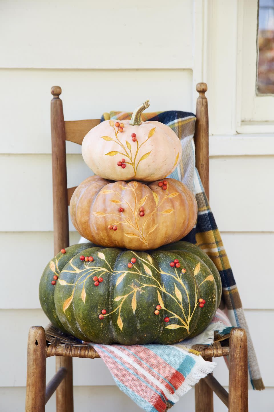 <p>The perfect sophisticated decor for greeting guests to your front door. Place directly on the porch or layer on a vintage ladderback chair with different color plaid blankets.</p><p><strong>To make:</strong> Purchase one large, one medium, and one small pumpkin (any color combo works) that stack nicely. Remove the stems from the large and medium pumpkin. Lightly sketch a vine pattern on a pumpkin with a pencil. Use a linoleum carving tool to etch out the pattern. Once complete attach red berries or beads with hot glue.</p>