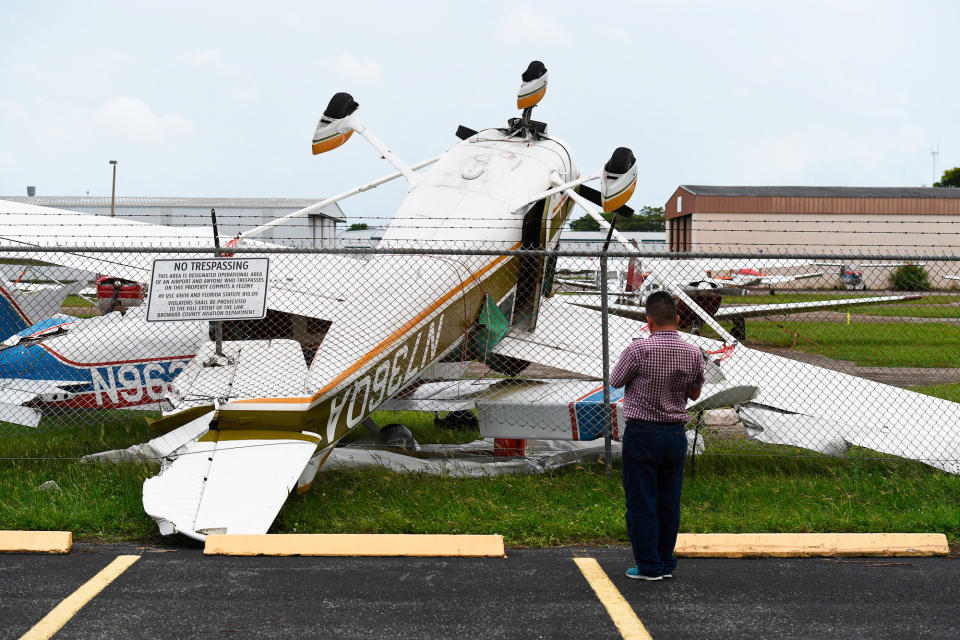 A person stands with their back to the camera; on the other side of chain-link fence from them are small destroyed planes in a heap