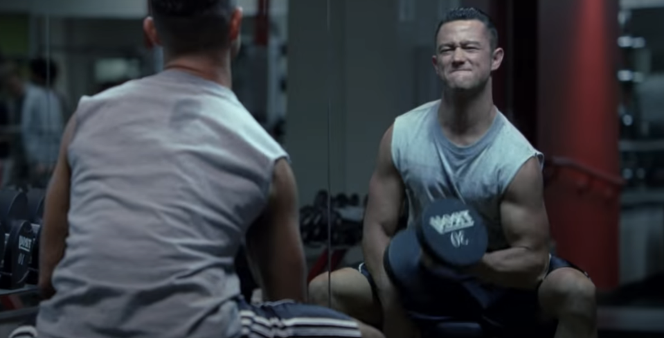 Joseph Gordon-Levitt is lifting a dumbbell with a determined expression in a gym, facing a mirror. He is wearing a sleeveless shirt and workout shorts