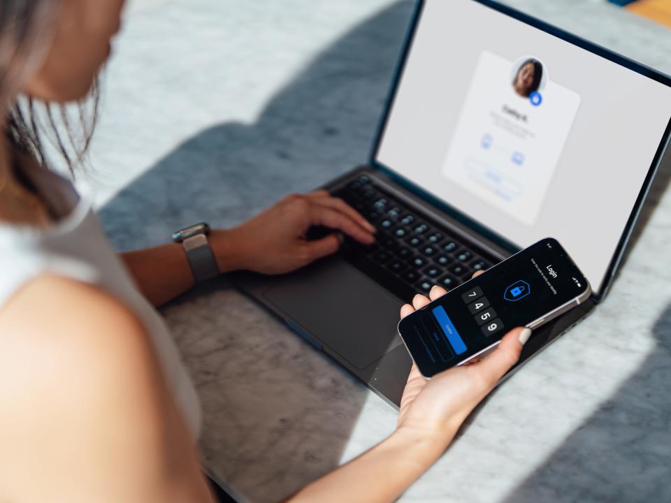 Over the shoulder view of young woman using smart phone while logging into an online account on laptop.