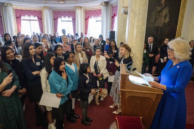 Camilla, wearing reading glasses, addresses a crowded room as she stands at a lectern in Buckingham Palace