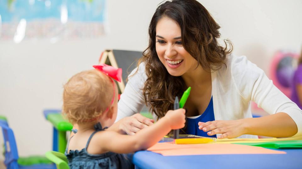 Beautiful Hispanic female daycare teacher leans on table and helps cute blonde toddler girl with school project.