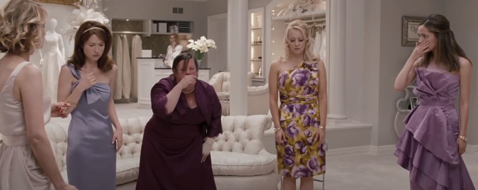 Five women in a bridal shop, expressing varied emotions, dressed in formal wear