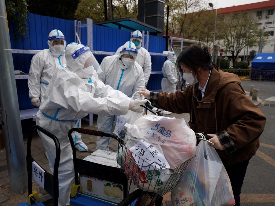 masked workers in white biohazard suits take bags of food from woman with cart