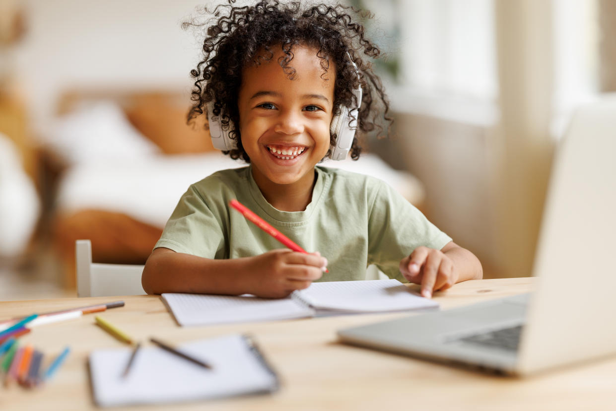 Distance education. Smiling african american child school boy in headphones studying online on laptop at home, sitting at table and communicating with teacher through video call on computer