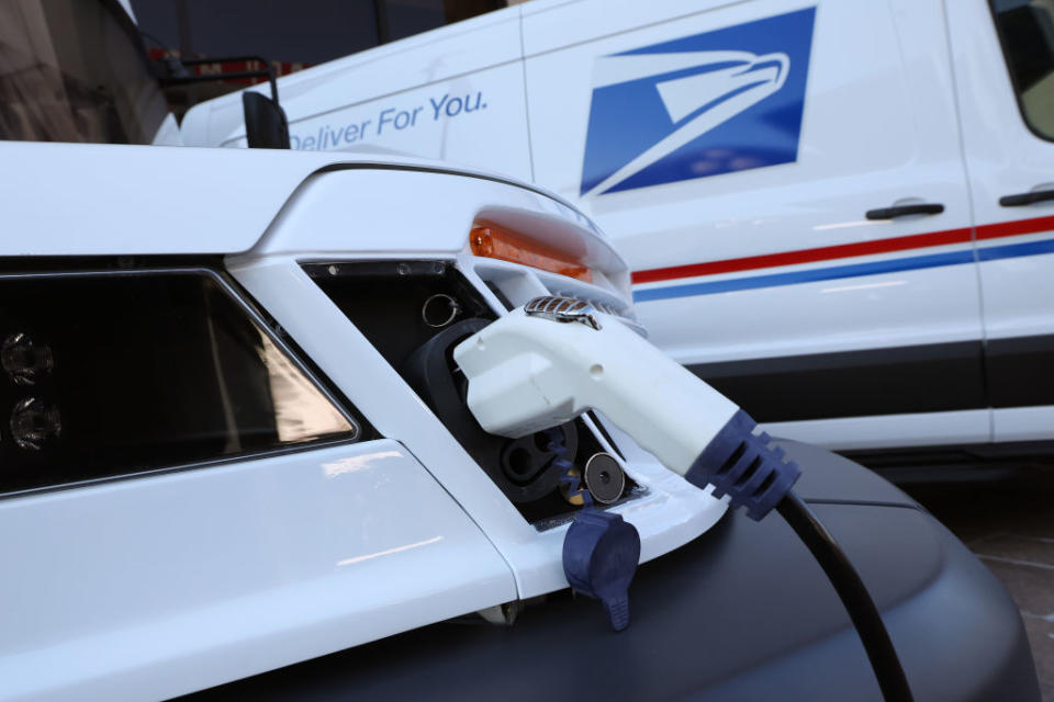 An electric vehicle is being charged with a postal service van in the background. The van bears the U.S. Postal Service logo and slogan 