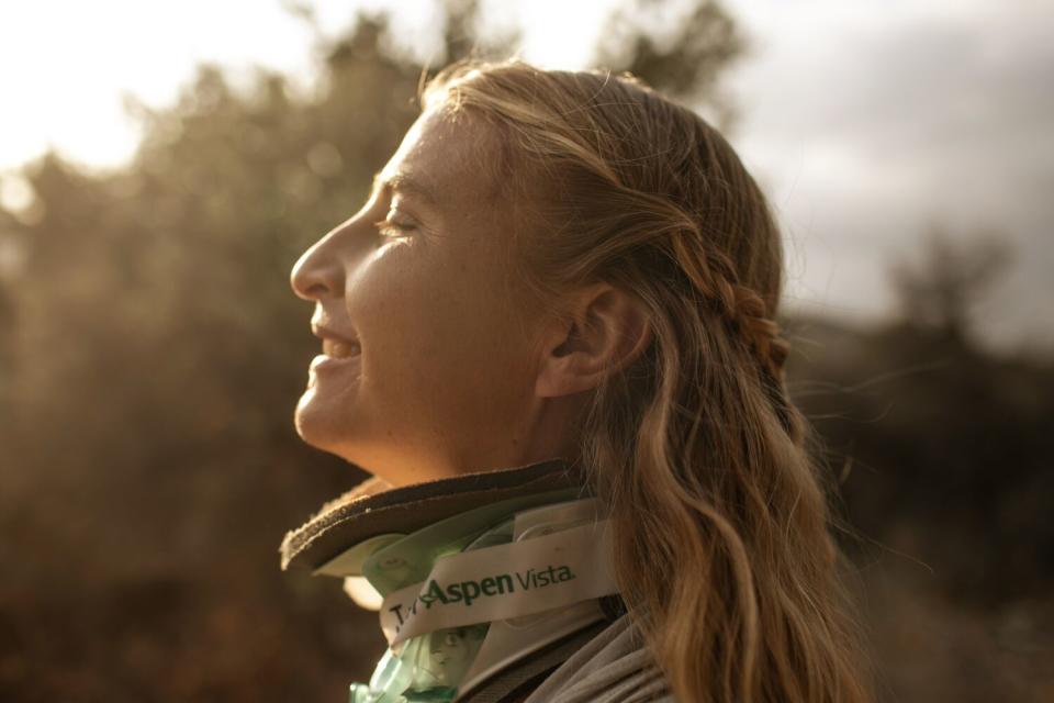 A blonde woman, in a head-and-shoulders profile view, lifts her head with eyes closed, smiling into sun-setting light.