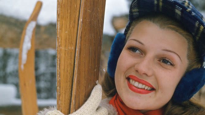 rita hayworth smiles and looks left of the camera, she wears a tan fleece jacket, orange scarf, and blue plaid hat and holds two wooden skis