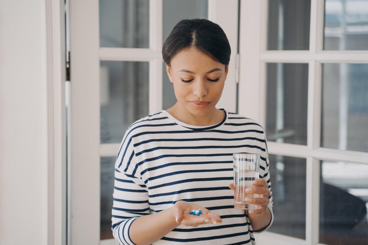 Pensive woman holds pills and glass of water doubting whether to take medicines. Thoughtful female has doubts about medications holding tablets in palm, standing at home. Healthcare, self-medicating.