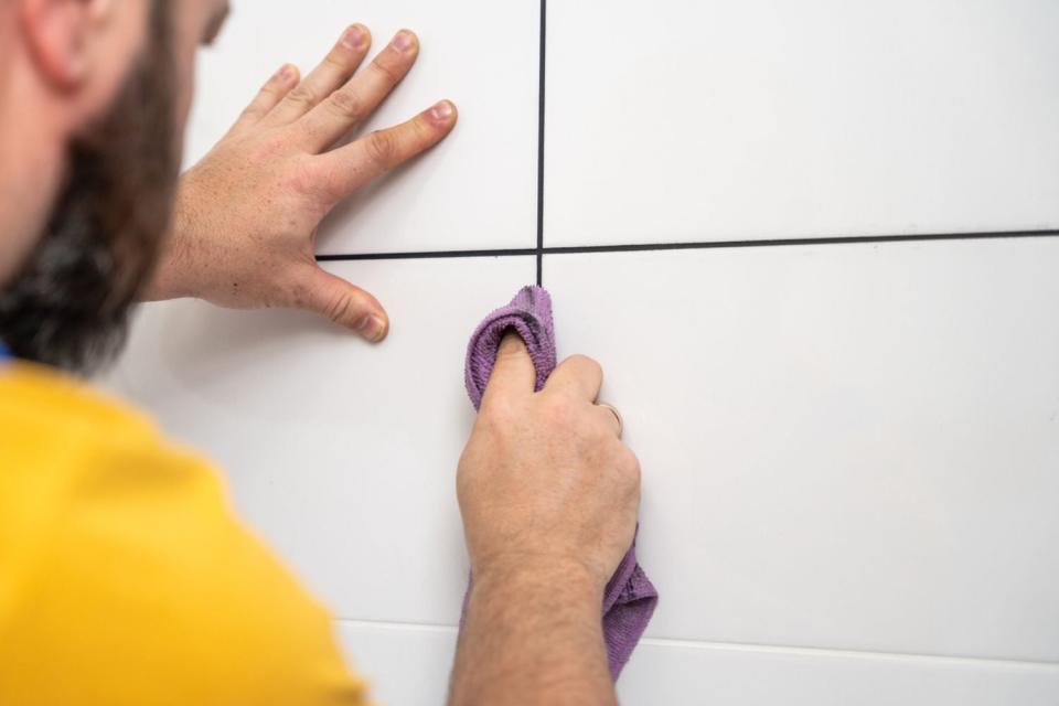 A worker in a yellow shirt uses a purple rage to clean the grout between white bathroom tiles. 