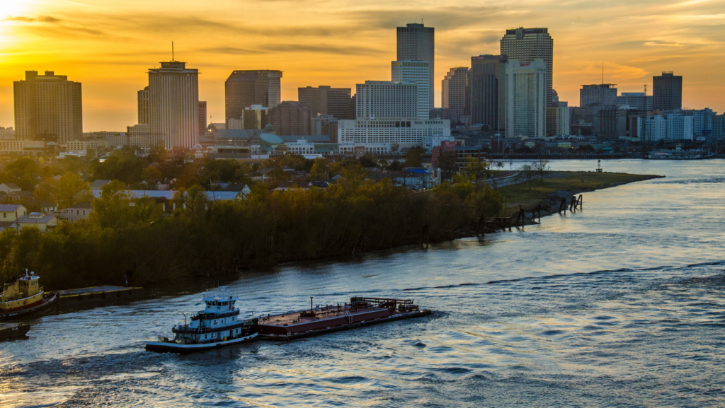 A tug boat pulls a barge on the Mississippi River downriver from the city of New Orleans, shown on the horizon at sunset