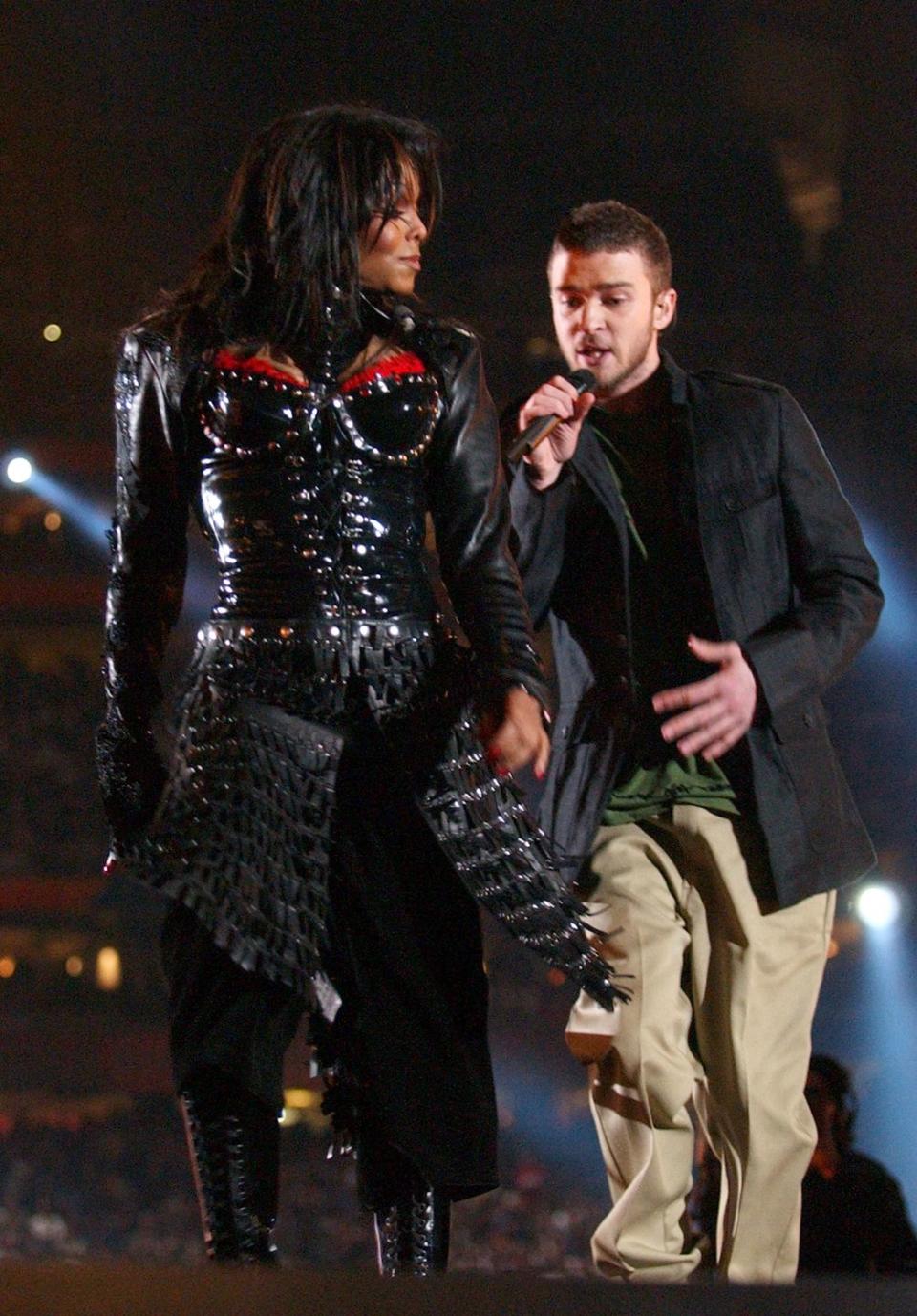 janet jackson and justin timberlake walk on a stage as he sings into a microphone, she wears a mostly black outfit, he wears a dark jacket over a green shirt and khaki pants