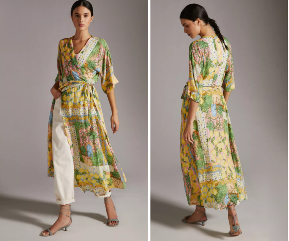 A black haired olive skinned woman wears a green and yellow mediterannean-patterned ankle length kimon wrap dress with 3/4 sleeves against a grey background.