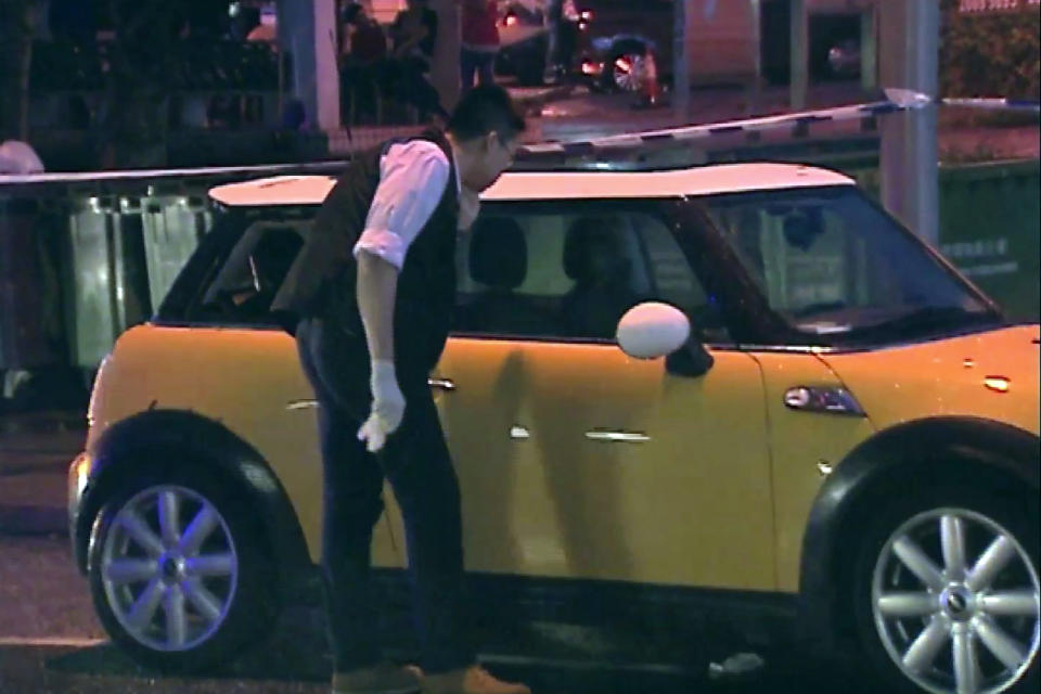 In this May 22, 2015, image from video provided by Now TV, a police officer searches and collects evidence in a yellow mini cooper where the suspect's wife and daughter were found unconscious in Hong Kong. An anesthesiologist is on trial in Hong Kong charged with killing his wife and daughter by placing a leaking yoga ball filled with carbon monoxide in their car. (Now TV via AP)
