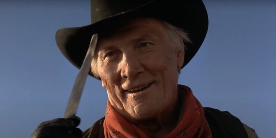 Man in cowboy attire with hat and neckerchief smiles on a clear day