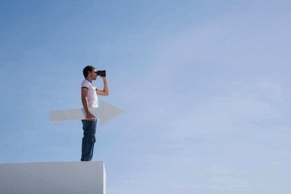 A person stands on a roof with an arrow and binoculars in his hand.