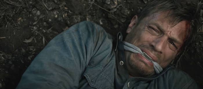 A man lies on the ground with a gag in his mouth, appearing distressed and injured, from what seems to be a dramatic scene in a film or TV show