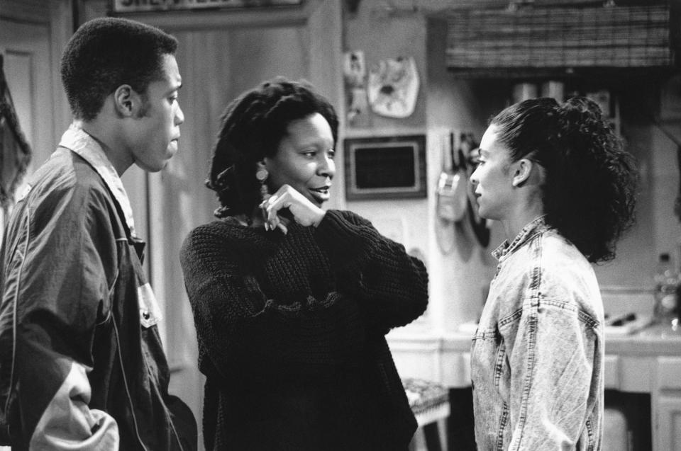 Whoopi Goldberg, standing between a man and a woman, engaged in conversation in a scene from a TV show. All three are in casual clothing