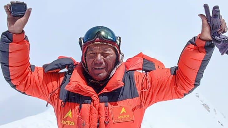 Dawa Ongju Sherpa on a summit, wearing red sit and goggles on his forehead, arms raised in celebration