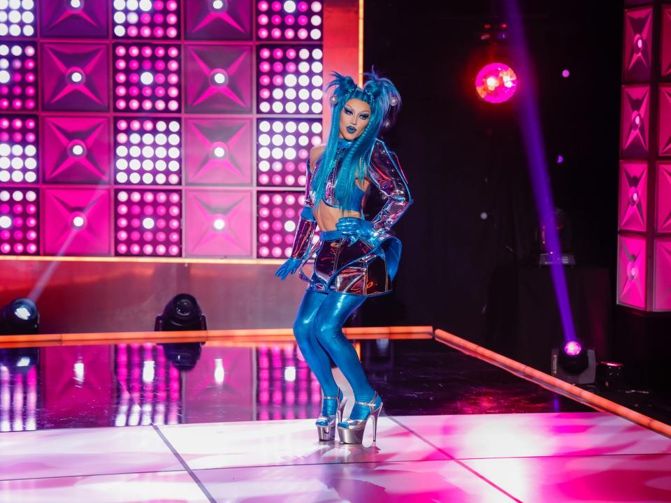 Spice poses on the runway in blue tights, silver heels, a purple mini skirt, and blue hair in this still from episode 3 of "RuPaul's Drag Race" season 15.