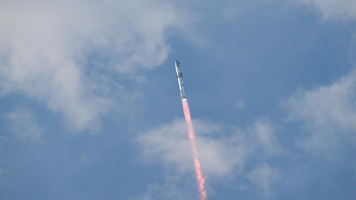  A large silver rocket flies through a blue sky above a column of flame. 