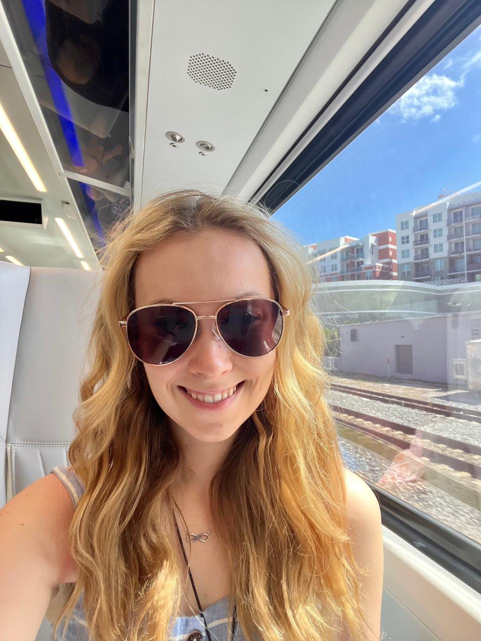 A woman wearing aviator sunglasses is shown inside a Brightline train car.