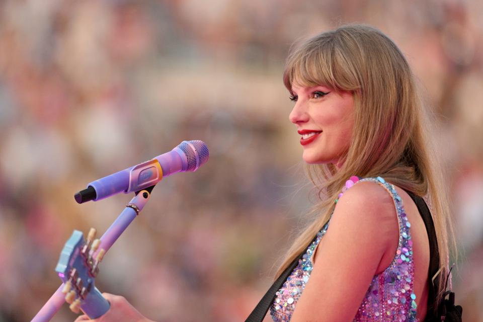 Taylor Swift wearing a sparkly crop top and smiling while playing a blue acoustic guitar and singing into a microphone onstage in front of a crowd.