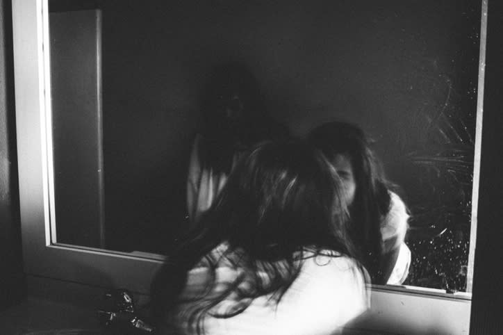 Person reflected in a mirror in a dimly lit room, with a contemplative posture