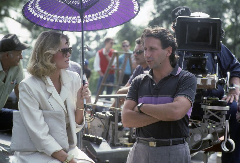 A woman with blonde hair in a white suit holding an umbrella speaking to a man on her right