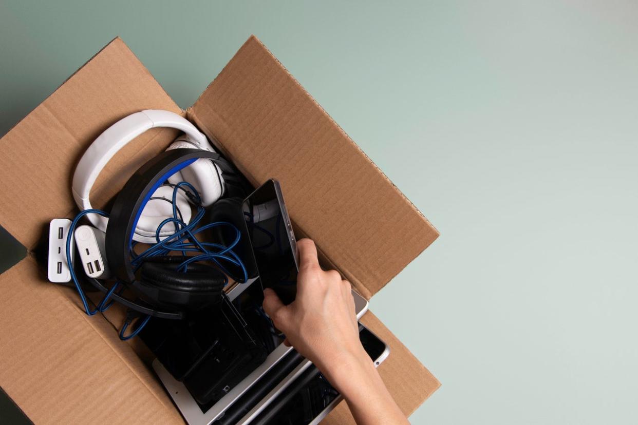 A woman's hand places a cell phone in a small cardboard box full of electronics for donation.