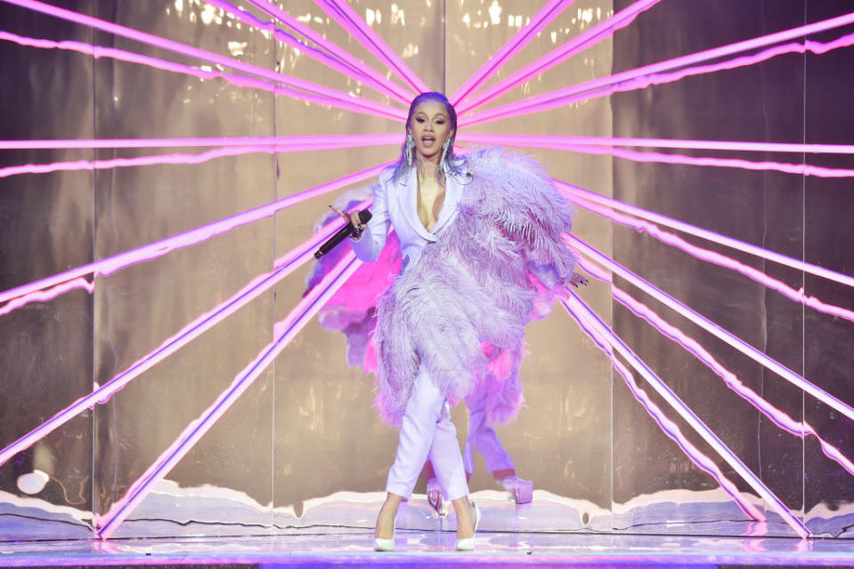 Cardi performing in a light-colored pantsuit with large feathers coming across her body