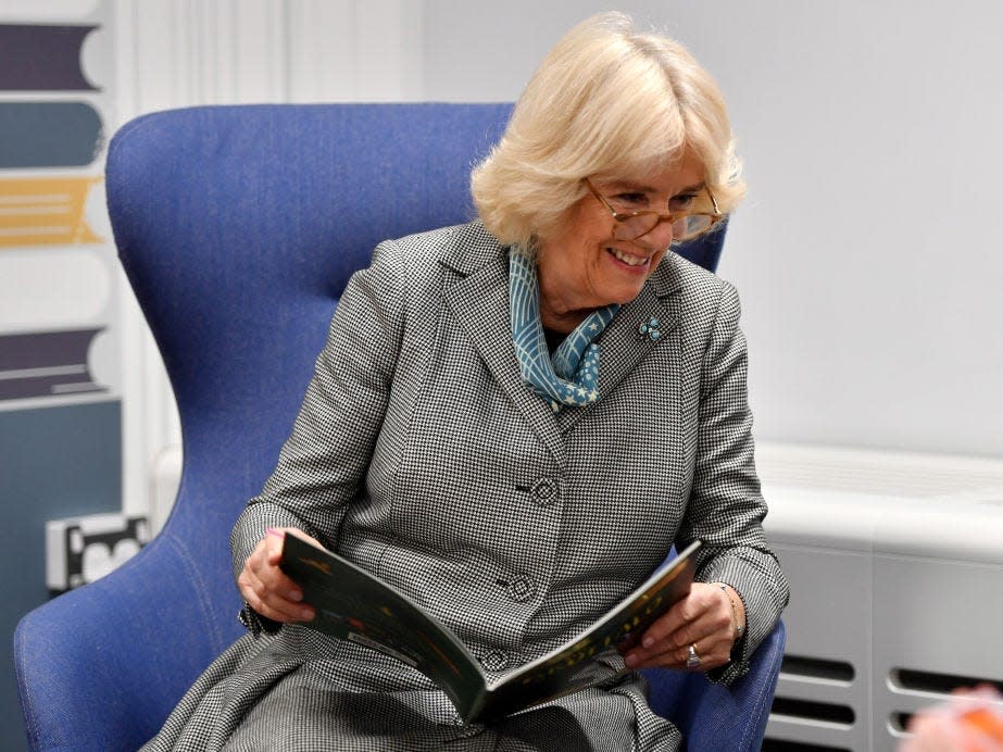 Camilla reads a book while sitting in a blue chair and smiling