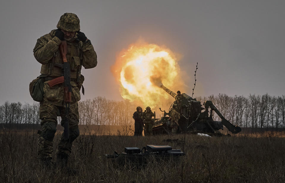 A helmeted Ukrainian soldier blocks his ears as a group of four fellow soldiers blast a missile from an armored vehicle, with an avenue of trees in the background. 