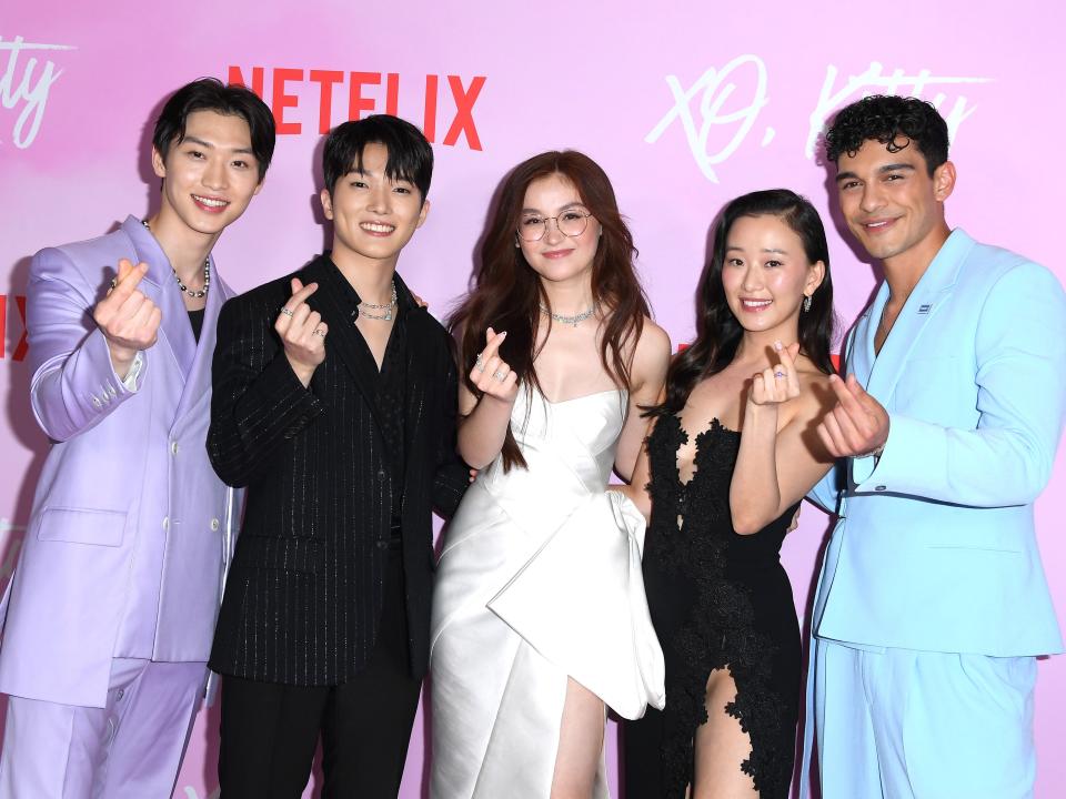 sang heon lee, minyeong choi, anna cathcart, gia kim, and anthony keyvan at the xo kitty premiere. they're all standing together on the red carpet: lee in a pastel purple suit, choi in a black suit, cathcart in a white gown, kim in a black gown, and keyvan in a pastel blue suit. they're all making finger hearts