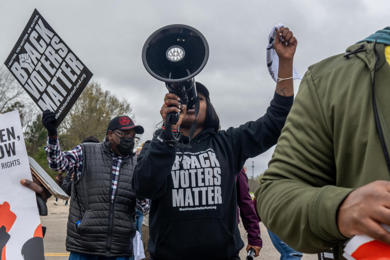 I'munique Liggens speaks into a loudspeaker, her left fist raised and wearing a sweatshirt that reads Black Voters Matter, as a fellow protester in a black COVID mask behind her waves a poster with the same slogan.