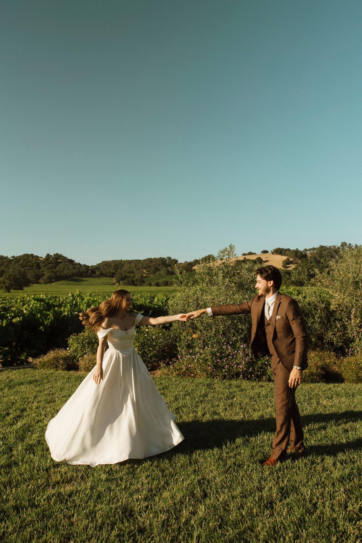 A bride and groom hold hands in a field in their wedding attire.