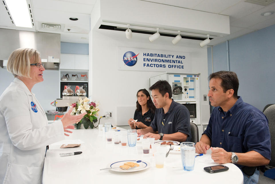 A woman in a white lab coat stands in front of three people in dark shirts sitting at a white table with food and drinks.