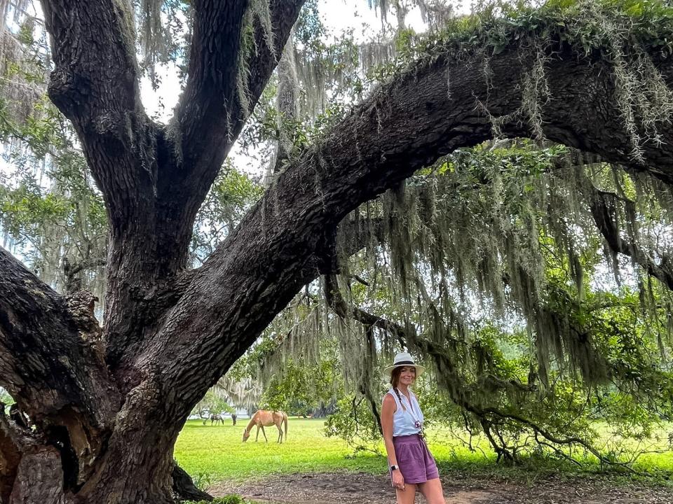 Emily standing under a large tree with moss hanging down. There is a brown horse in the background.